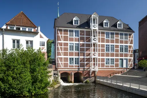 Au&szlig;enansicht des Praxisstandorts in Hamburg-Bergedorf bei sonnigem Wetter: Ein historisches, vierst&ouml;ckiges Fachwerkhaus mit roter Backsteinf&uuml;llung steht unmittelbar am Wasser des Eppendorfer M&uuml;hlenteichs. Rechts f&uuml;hren Steinstufen zum Wasser hinunter