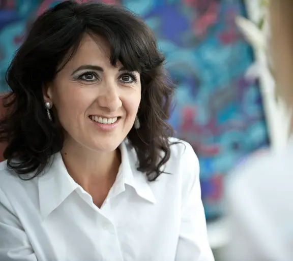 Portr&auml;t von Prof. Dr. med. Alaleh Raji. Die &Auml;rztin hat dunkles, schulterlanges lockiges Haar und blickt mit einem freundlichen, offenen L&auml;cheln leicht zur Seite. Sie tr&auml;gt eine wei&szlig;e Hemdbluse und dezente Ohrringe; der Hintergrund zeigt ein abstraktes Kun