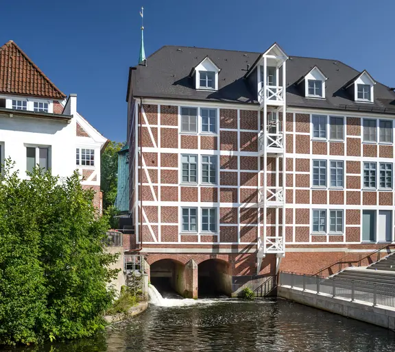 Au&szlig;enansicht des Praxisstandorts in Hamburg-Bergedorf bei sonnigem Wetter: Ein historisches, vierst&ouml;ckiges Fachwerkhaus mit roter Backsteinf&uuml;llung steht unmittelbar am Wasser des Eppendorfer M&uuml;hlenteichs. Rechts f&uuml;hren Steinstufen zum Wasser hinunter