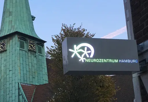 Nahaufnahme des beleuchteten Praxisschilds vom Neurozentrum Hamburg, das an einer Backsteinfassade montiert ist. Im Hintergrund ragt der gr&uuml;ne, kupferbeschlagene Turm der St. Johannis Kirche vor einem blauen Himmel auf.